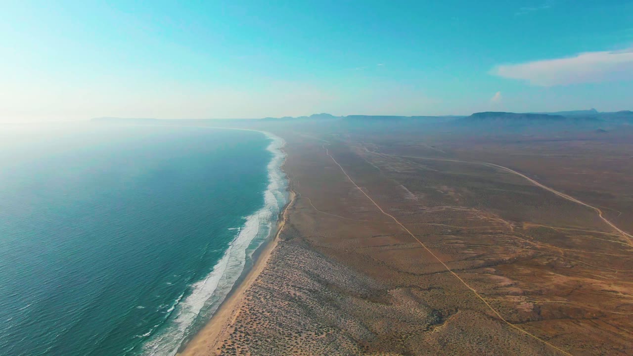 Aerial view over La Bocana with white waves angled along the beach, dry brown terrain spreading inland, a pale cloud over the far horizon, and no visible people.