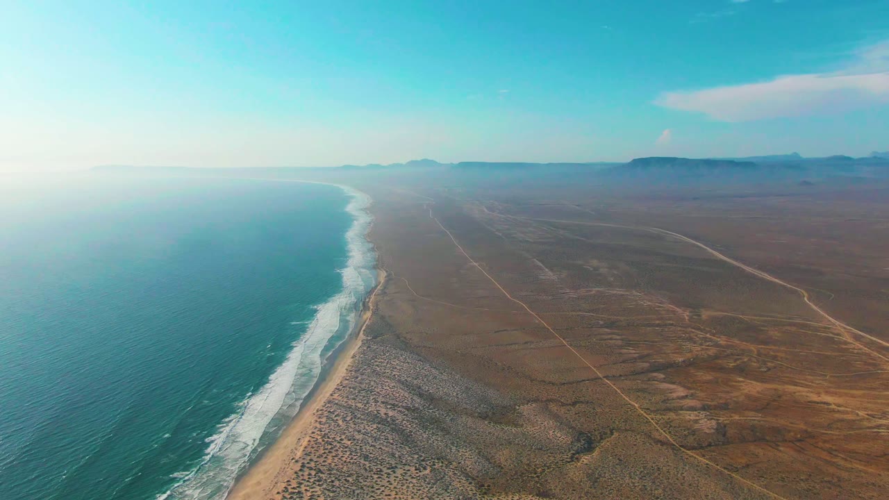 Aerial frame over La Bocana, Baja California Sur with turquoise ocean on the left, white surf tracing a sandy beach, brown desert terrain to the right, distant hazy hills, and no visible people.