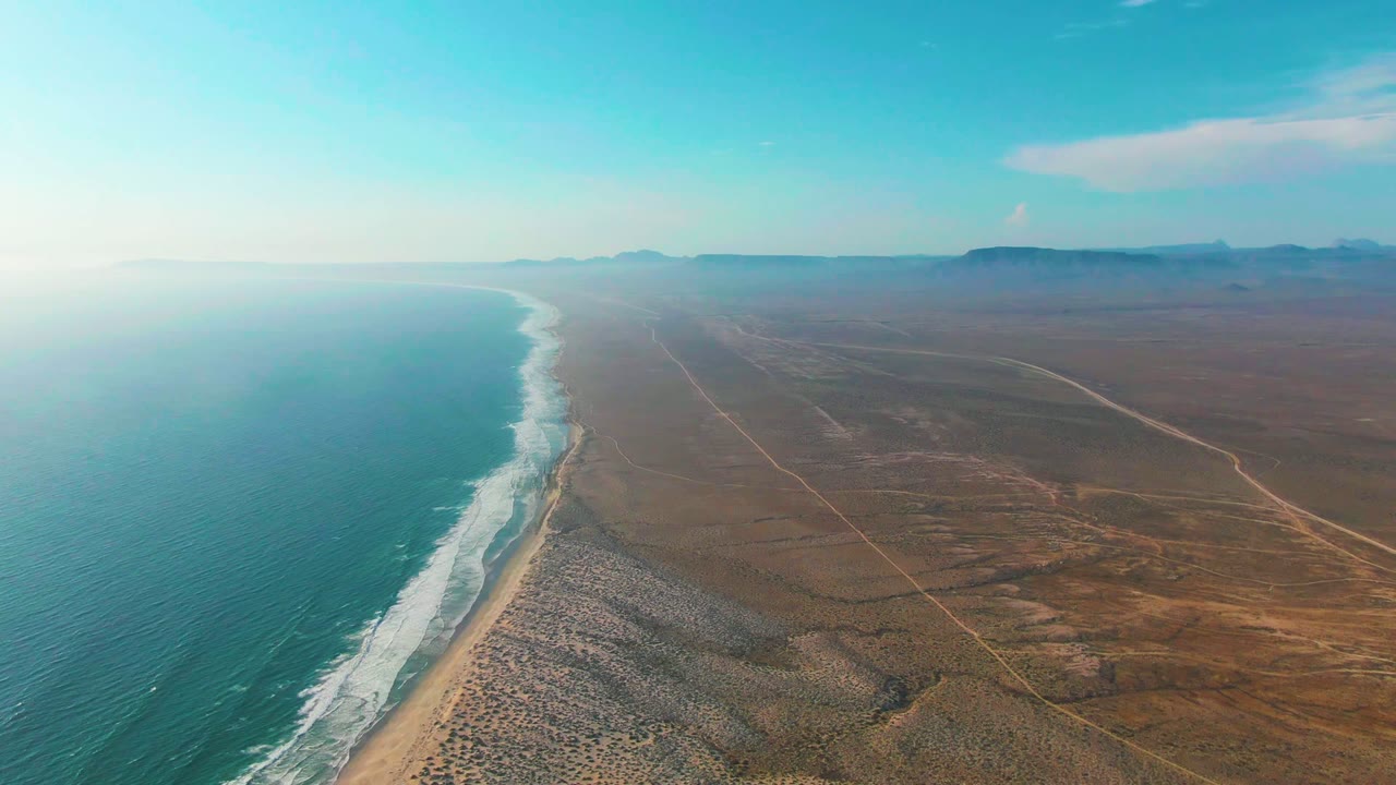 The La Bocana coastline remains centered with white wave bands along the beach, open turquoise water at left, faint dirt roads crossing the brown desert inland, distant mountains, and no visible people.