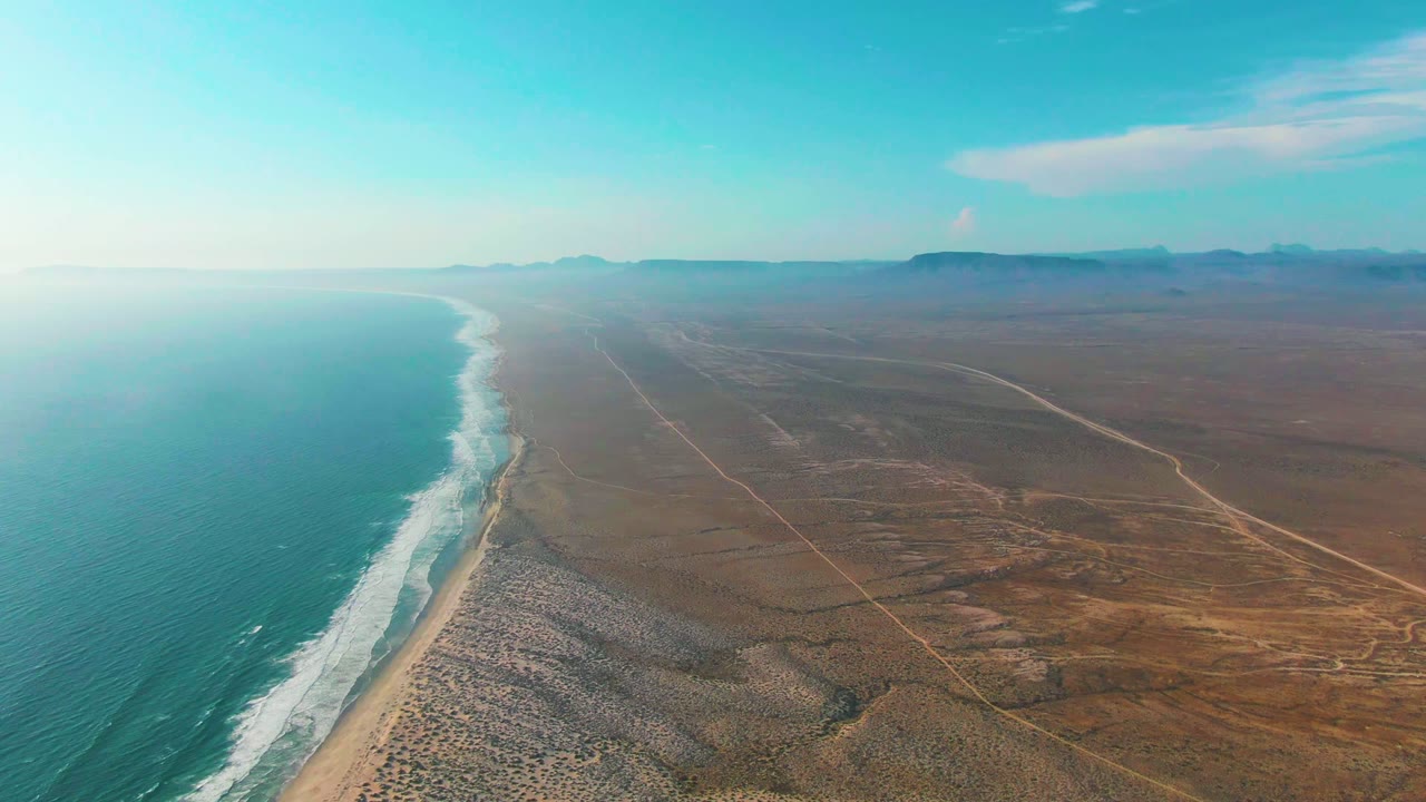 Opening aerial view of La Bocana, Baja California Sur, with turquoise ocean on the left, white surf along the sand, brown desert terrain to the right, distant hills, and no visible people.