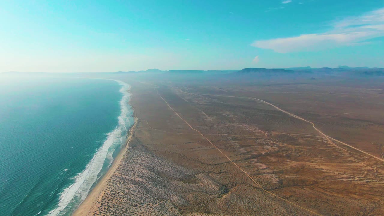 Aerial view of surf tracing a sandy shore beside open desert tracks.