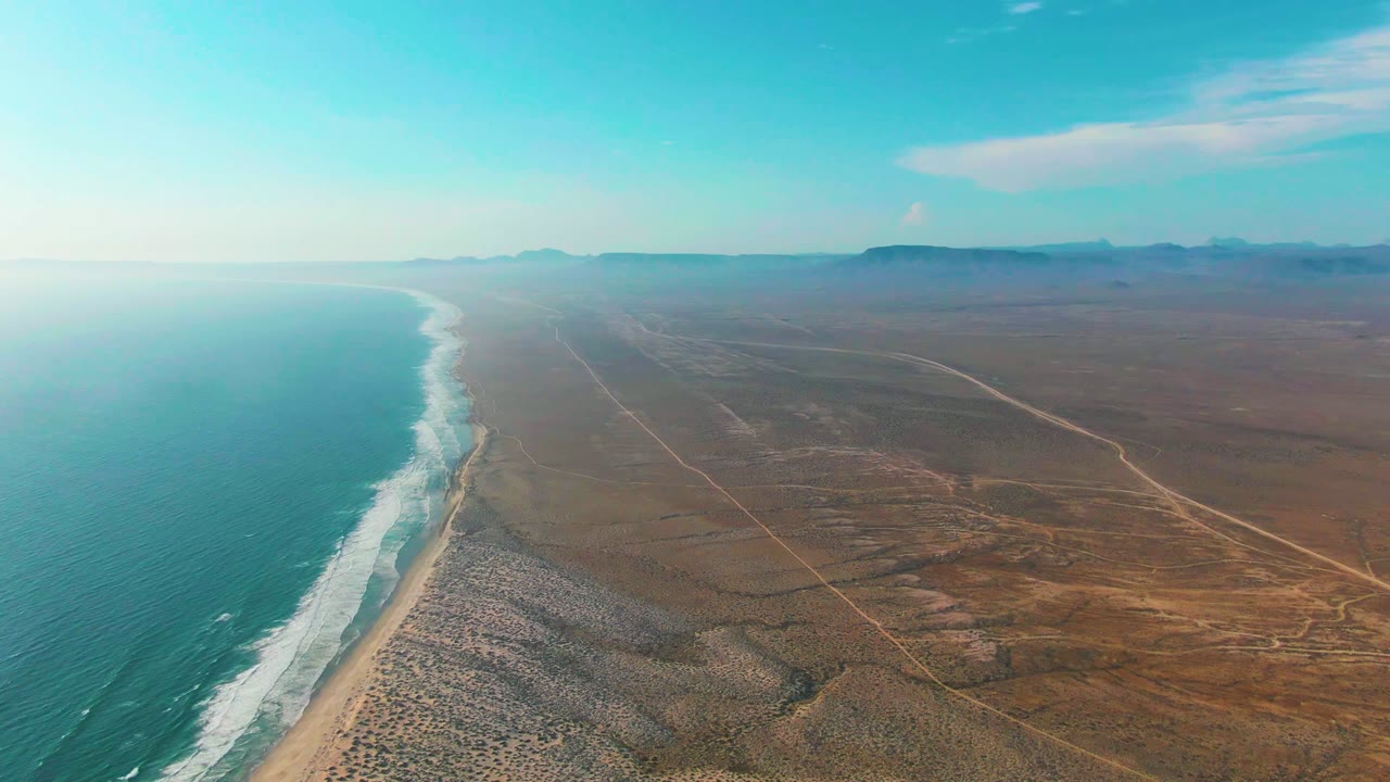 Aerial view over La Bocana with blue water and white waves on the left, a narrow sand strip, darker scrub patches across the desert, faint roads, distant mountains, and no visible people.