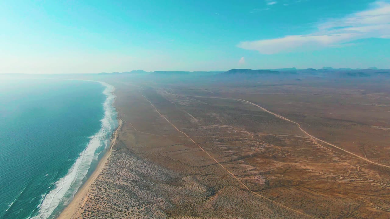 A high aerial view of La Bocana, Baja California Sur, shows turquoise ocean at left, a white surf line on the beach, brown desert flats to the right, distant mountains, and no visible people.