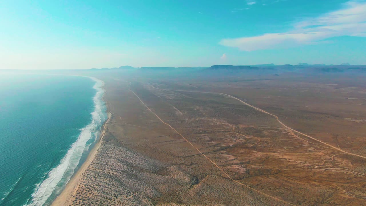 Aerial view of surf along a sandy beach beside open desert and road tracks.