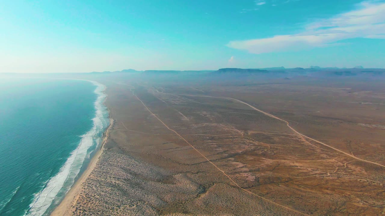 A wide drone-style frame over La Bocana shows the ocean pressed to the left edge, surf bands on the beach, a broad brown desert plain with crossing tracks, distant mountains, and no visible people.