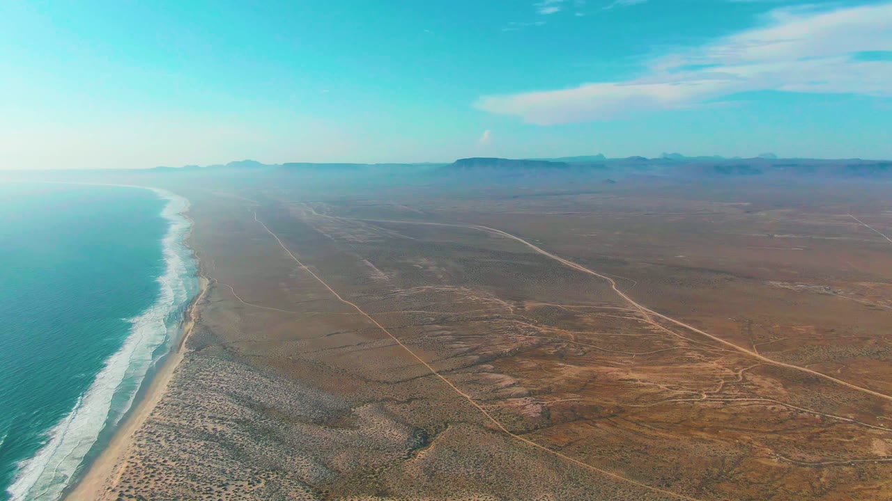 Aerial view over La Bocana, Baja California Sur, with turquoise ocean and white surf on the left, sandy beach and dry desert roads inland, and no visible people.
