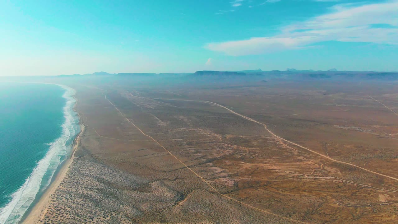 Aerial view over La Bocana with ocean waves rolling onto the beach at left, intersecting dirt roads across dry terrain at right, low distant landforms, and no visible people.