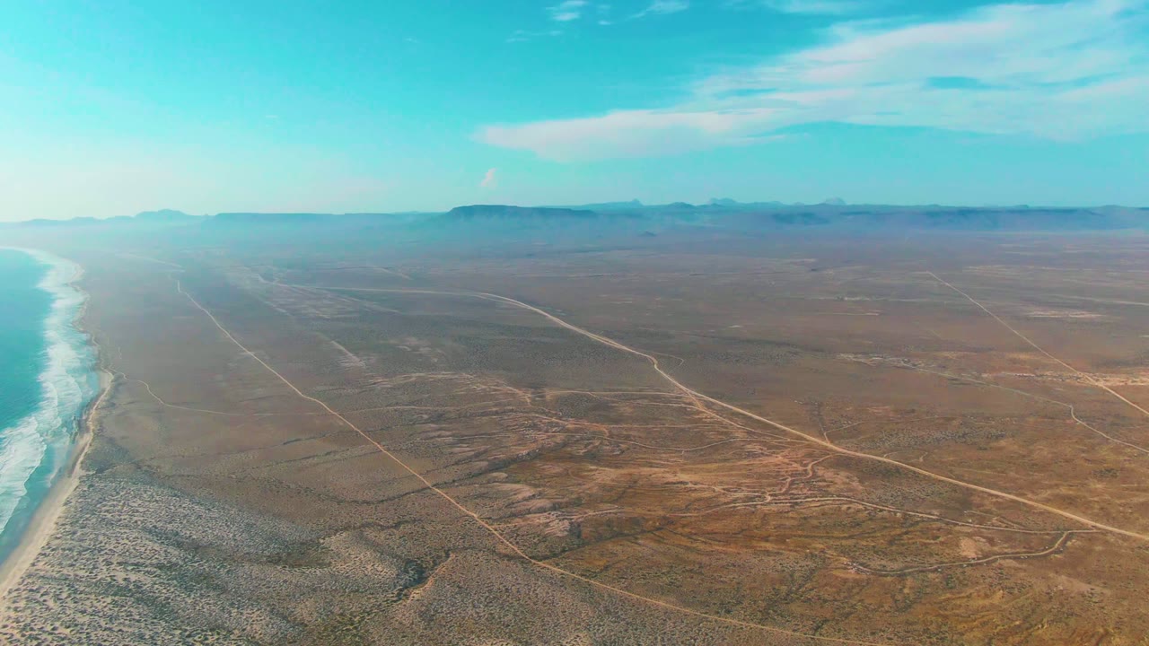 Aerial view over La Bocana, Baja California Sur with turquoise ocean and white surf on the left, brown desert roads inland, distant mountains, blue sky, and no visible people.