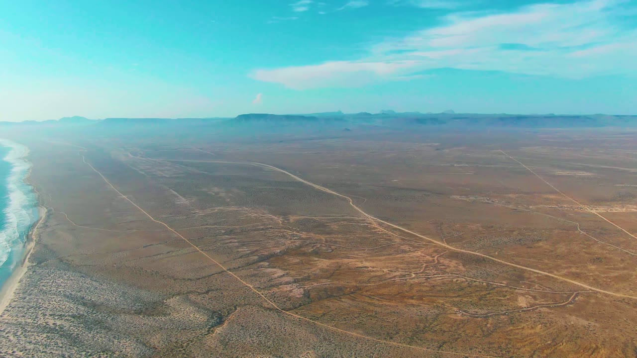 High aerial frame at La Bocana showing white surf against the left shoreline, a broad brown desert plain with intersecting roads, low mountains in the distance, and no visible people.