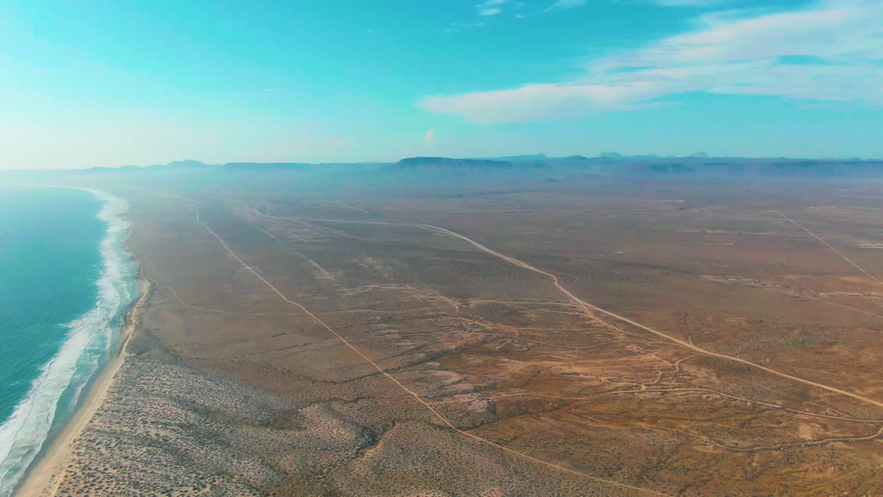 Aerial view over La Bocana with turquoise ocean and white surf at left, sandy shore below, brown desert roads inland, distant mountains, and no visible people.