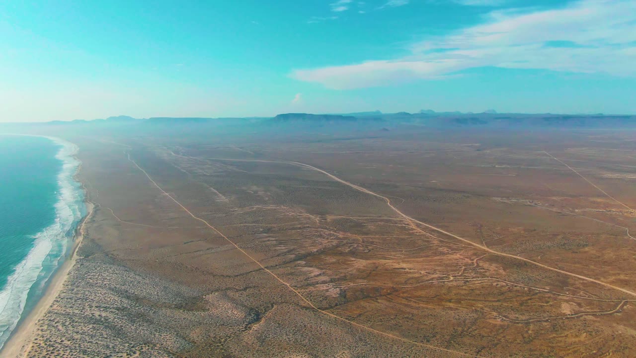 Aerial view of La Bocana with bright ocean water at far left, foamy waves, a narrow sandy beach, broad brown desert plain, faint roads, low mountains, and no visible people.