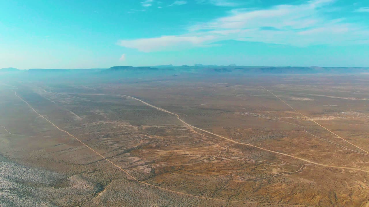 A high aerial view over La Bocana, Baja California Sur shows dry desert scrub, pale dirt roads crossing the foreground, distant low mountains, blue sky, and no visible people.