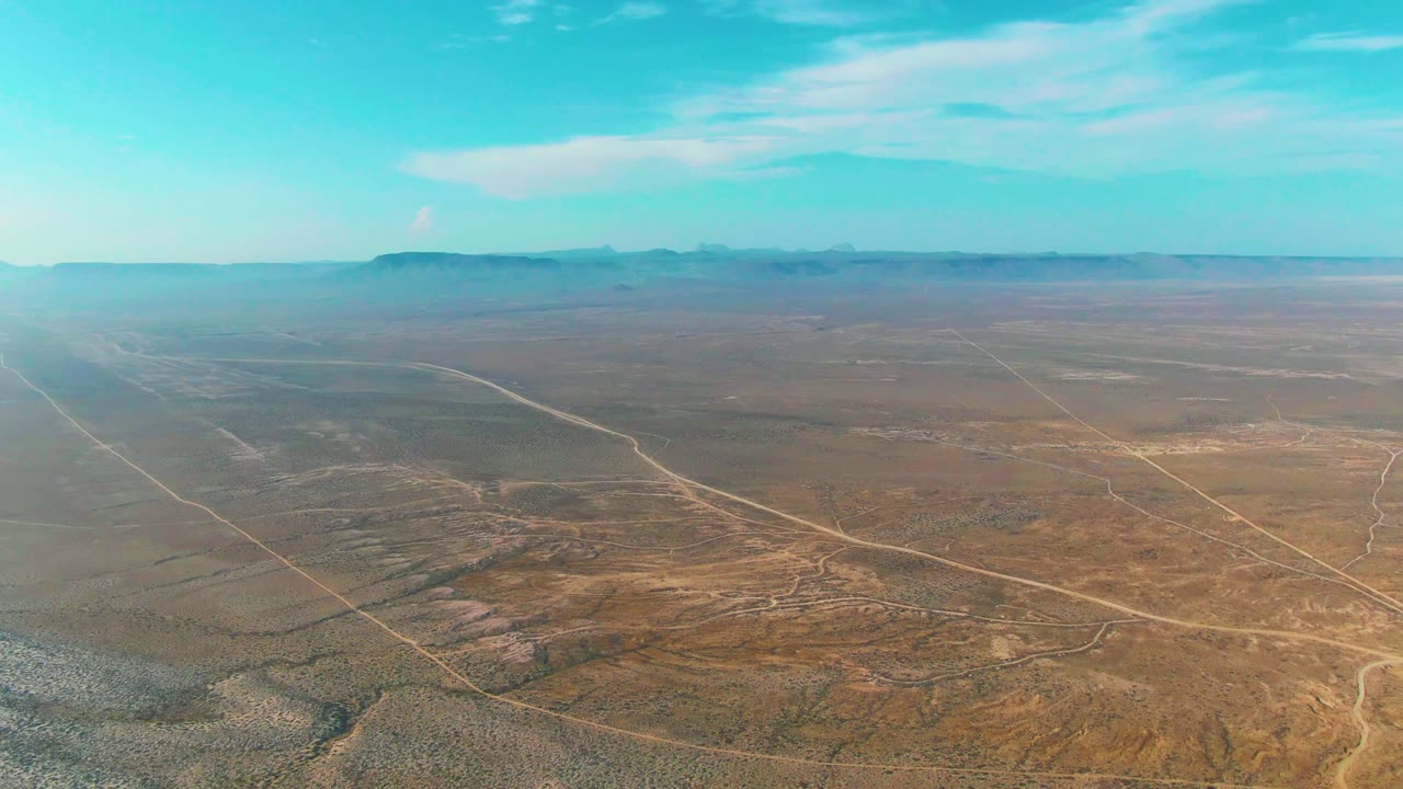 A drone-like aerial frame shows La Bocana desert terrain with a diagonal dirt road through the foreground, lighter road lines in the distance, low mountains, and no visible people.