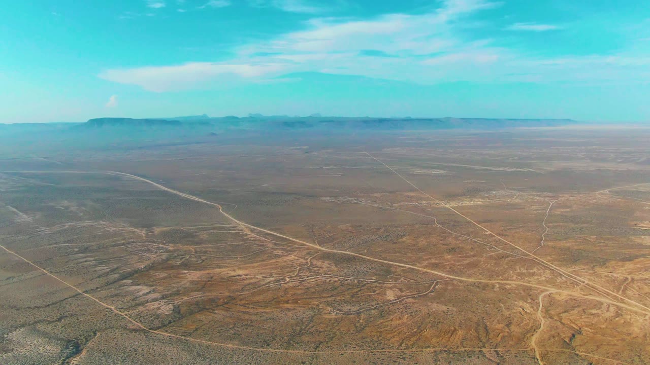 Aerial view over dry desert terrain near La Bocana, Baja California Sur, with pale dirt roads crossing the foreground, mountains on the horizon, blue sky above, and no visible people.