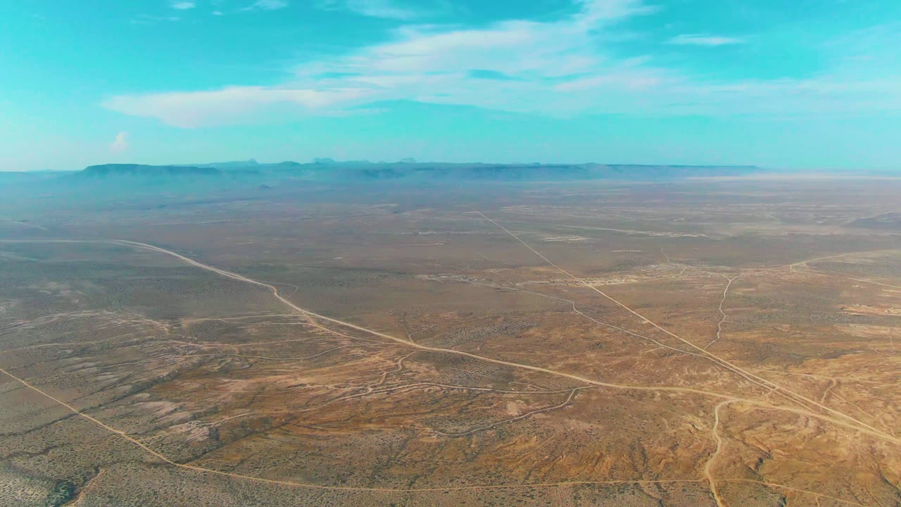 Aerial view of dry Baja California Sur terrain where several dirt roads and faint tracks stretch across the midground toward a flat, distant mountain horizon, with no visible people.