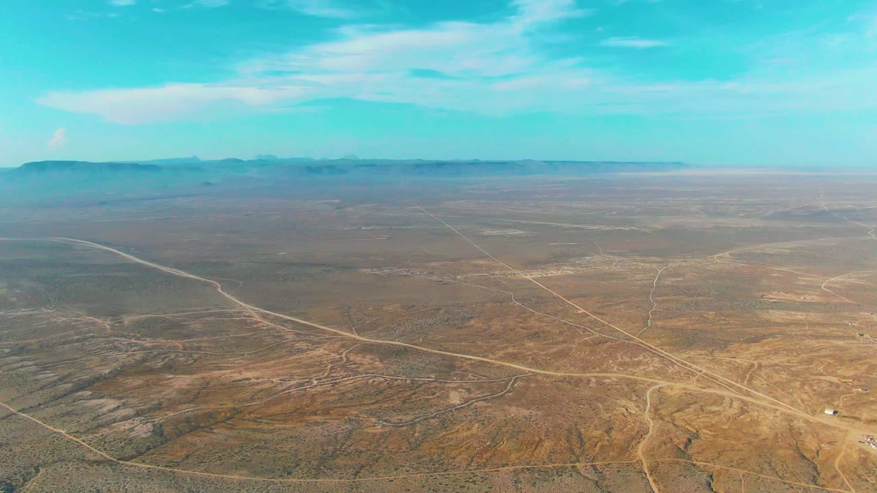 Wide aerial frame over La Bocana, Baja California Sur desert terrain with pale dirt roads, distant mountains, thin clouds, and no visible people.