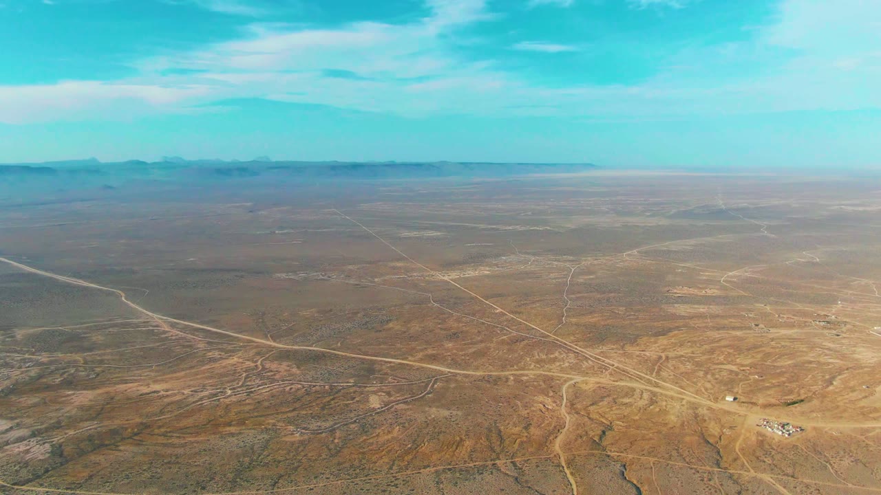 Aerial frame over La Bocana, Baja California Sur showing broad brown desert plains, intersecting pale dirt roads, distant mountains, thin clouds, and no visible people.