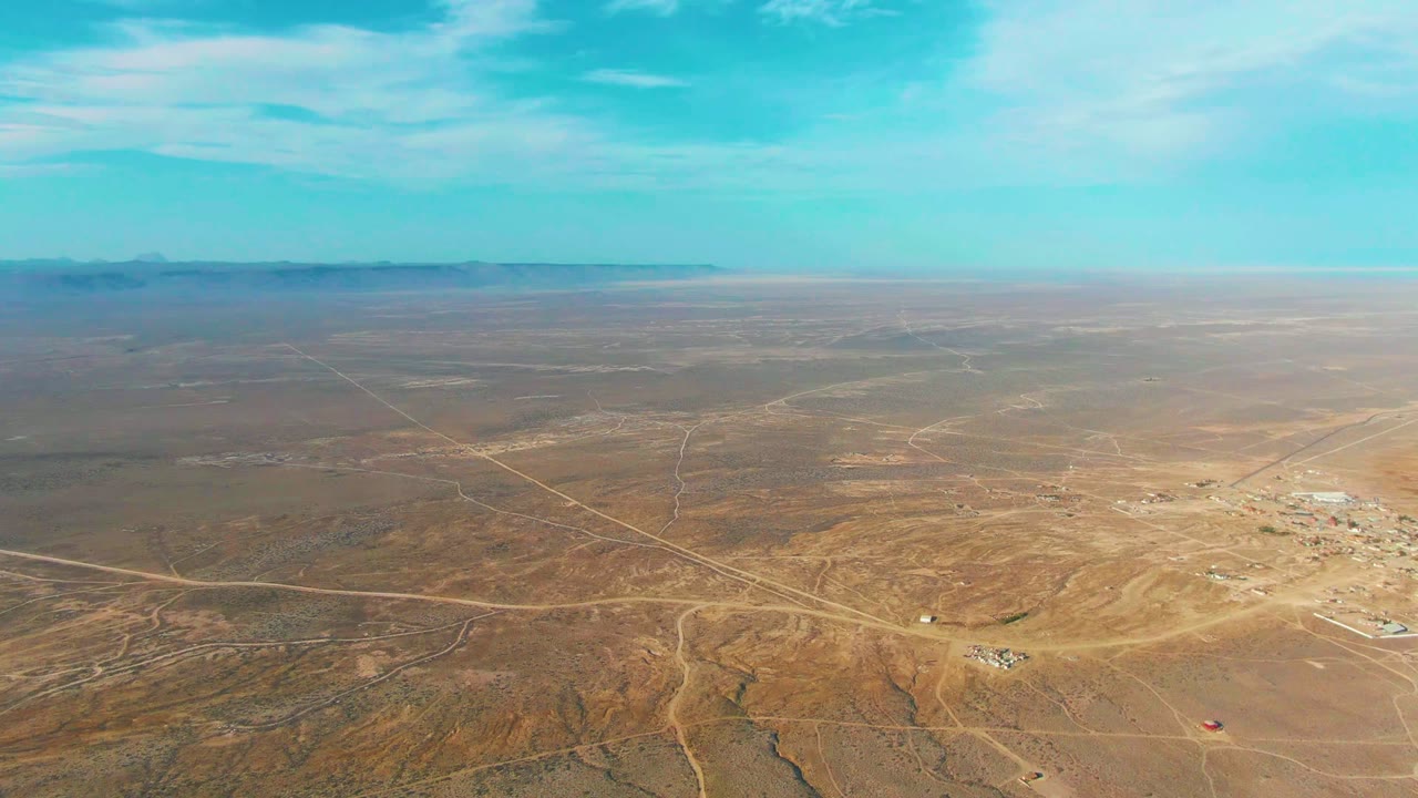 Wide aerial view over La Bocana, Baja California Sur desert terrain with intersecting dirt roads, small structures at right, distant low mountains, blue sky, and no visible people.
