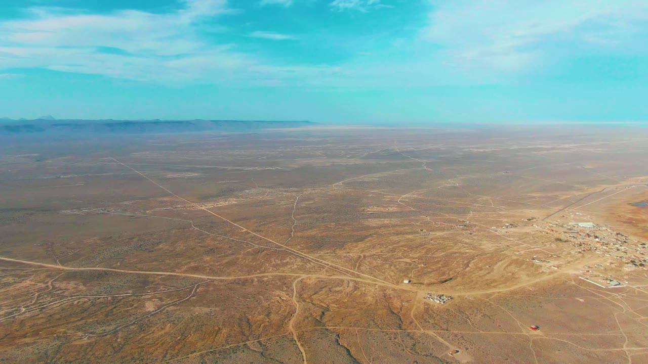 Drone-height view across the La Bocana desert, showing pale dirt roads, darker dry channels in the foreground, small structures to the right, a flat distant horizon, and no visible people.