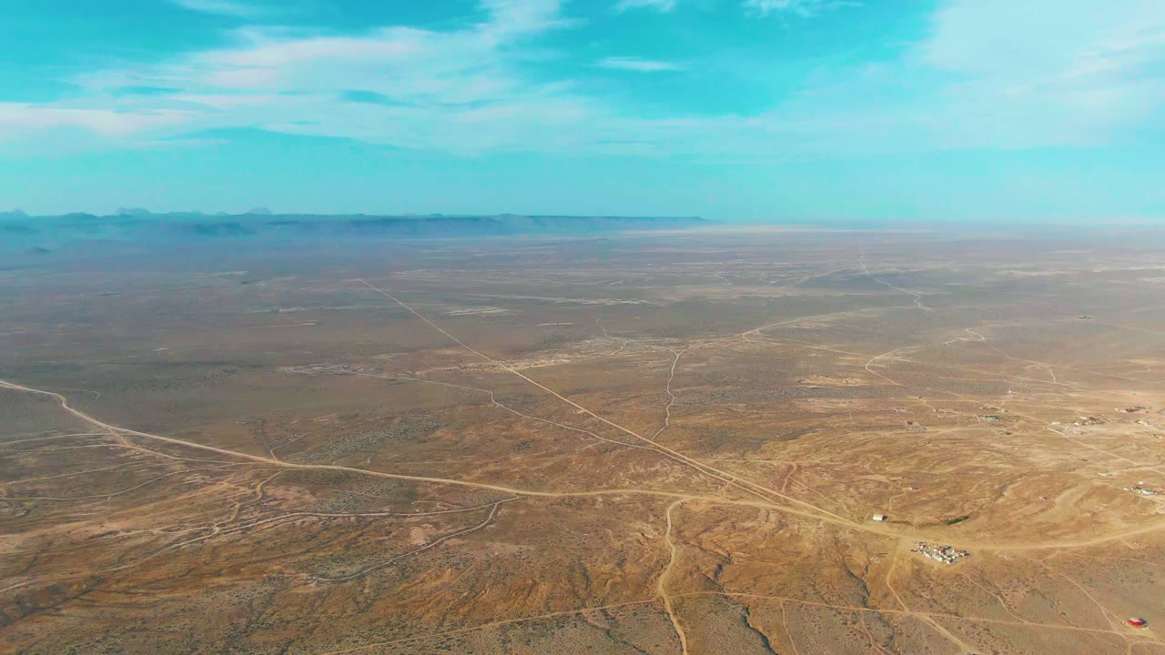 Opening aerial frame over La Bocana, Baja California Sur desert with pale dirt roads crossing dry brown terrain, distant mountains, blue sky, and no visible people.