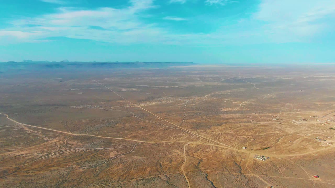 La Bocana aerial frame with dry sandy terrain, intersecting dirt roads, small structures near the lower right area, distant mountains, and no visible people.
