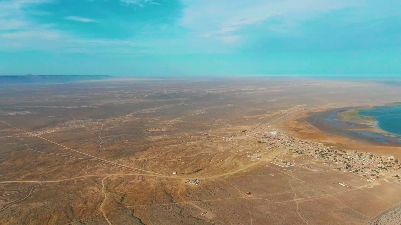 Aerial view over La Bocana, Baja California Sur, showing dry desert tracks, a compact settlement near blue water, a low horizon, and no visible people.