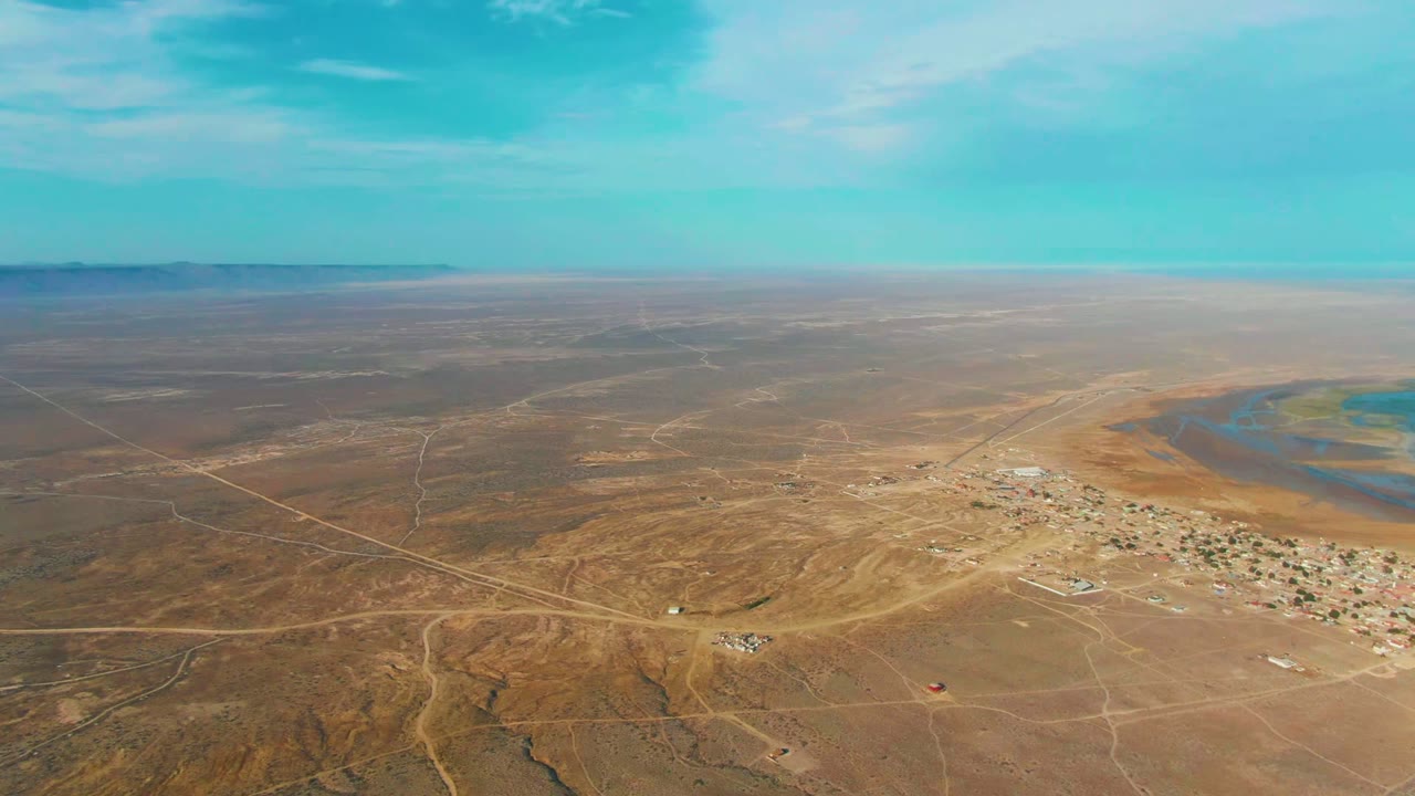 Wide aerial view over La Bocana, Baja California Sur, with brown desert terrain, a small settlement near blue water, distant mesas, and no visible people.