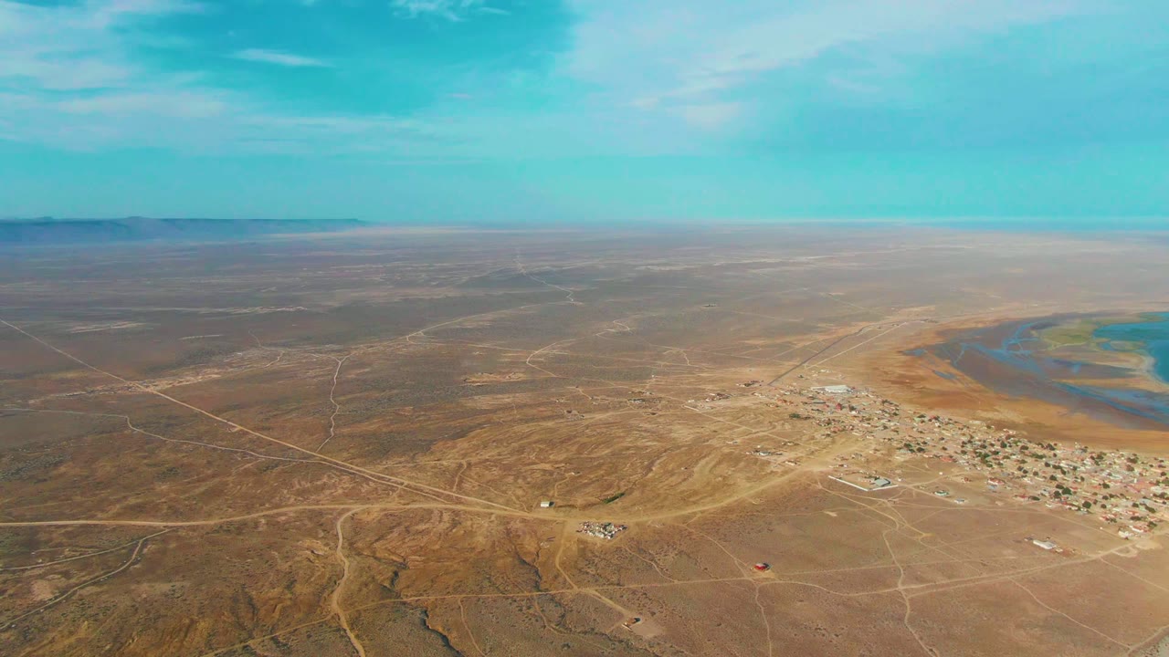 La Bocana appears from a high drone angle with scattered buildings, tan desert roads, a curved blue water area at right, a low horizon, and no visible people.