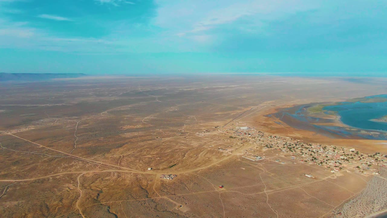 Wide aerial view over La Bocana, Baja California Sur, with brown desert terrain, a small settlement near shallow blue water, pale dirt roads, distant horizon, and no visible people.