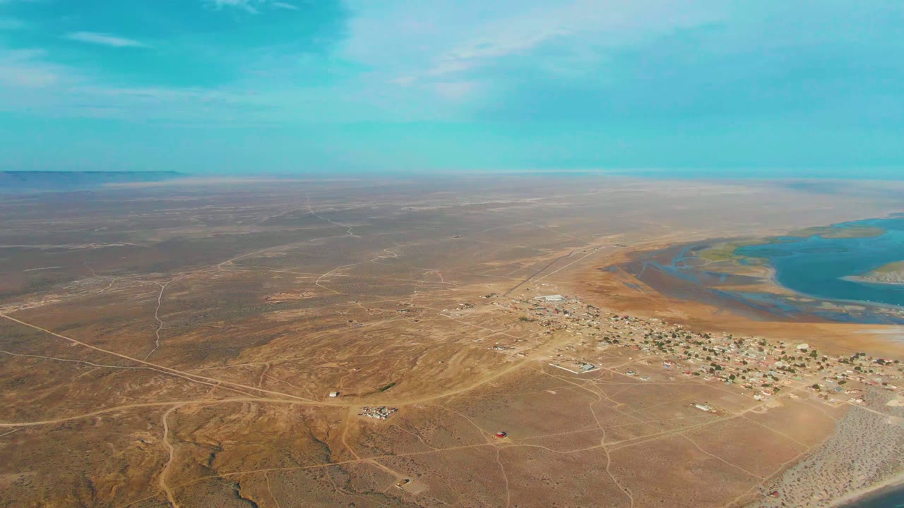 Aerial view over La Bocana with pale desert roads crossing the foreground, clustered camp buildings in the midground, turquoise shallow water on the right, and no visible people.