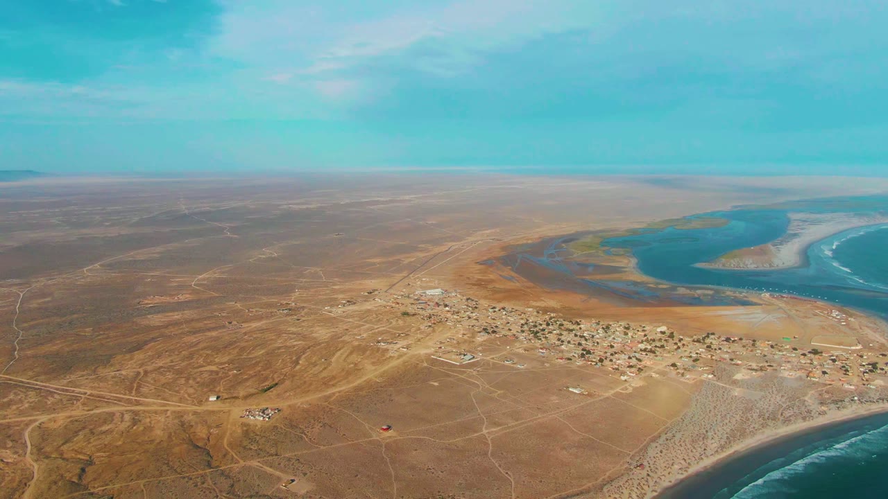 Wide aerial view over La Bocana, Baja California Sur, with arid brown terrain, a small settlement near the shore, blue estuary water, ocean surf, and no visible people.