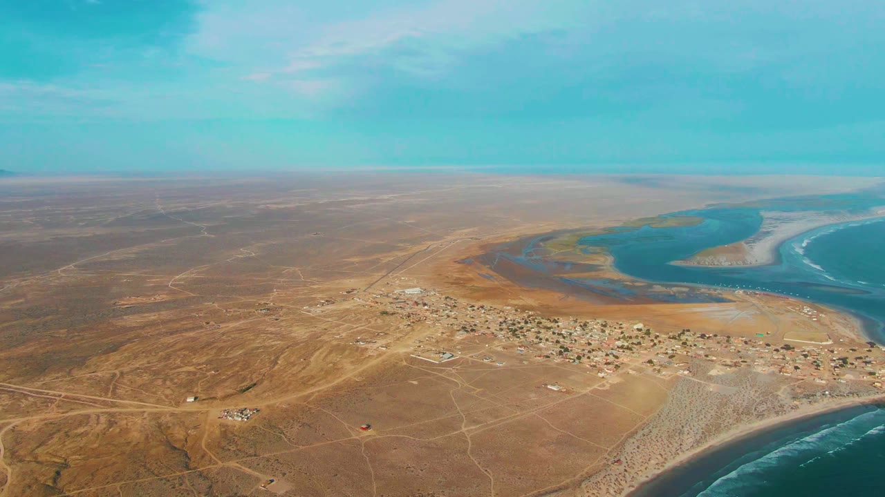 High aerial view of La Bocana with tan desert land filling the foreground, scattered settlement buildings near the shore, blue estuary water behind them, breaking ocean waves, and no visible people.