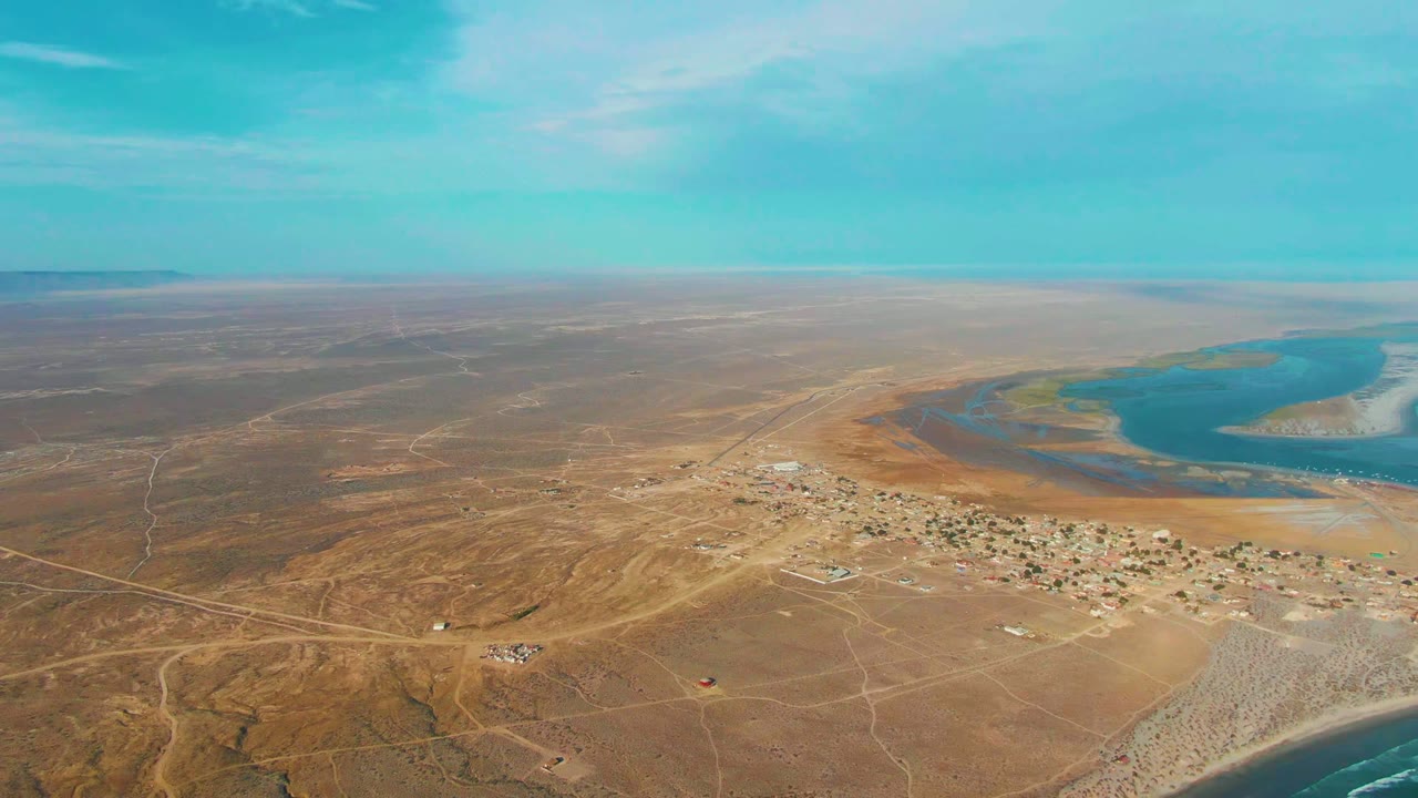 Wide aerial frame over La Bocana, Baja California Sur, showing dry brown terrain, a small settlement, pale roads, blue inlet water, ocean surf at the lower edge, and no visible people.