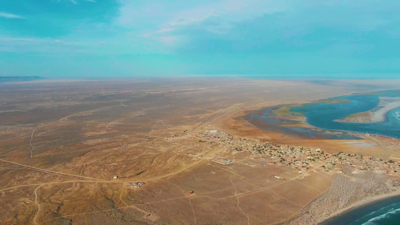 Aerial frame over La Bocana, Baja California Sur, with brown desert foreground, scattered buildings in the midground, lagoon-colored water to the right, a distant flat horizon, and no visible people.