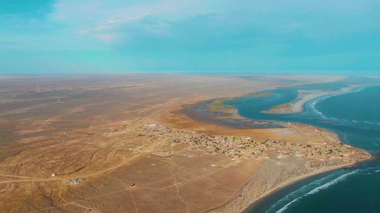 High aerial view over La Bocana, Baja California Sur, showing a small coastal settlement, brown desert terrain, a winding blue waterway, ocean surf, and no visible people.