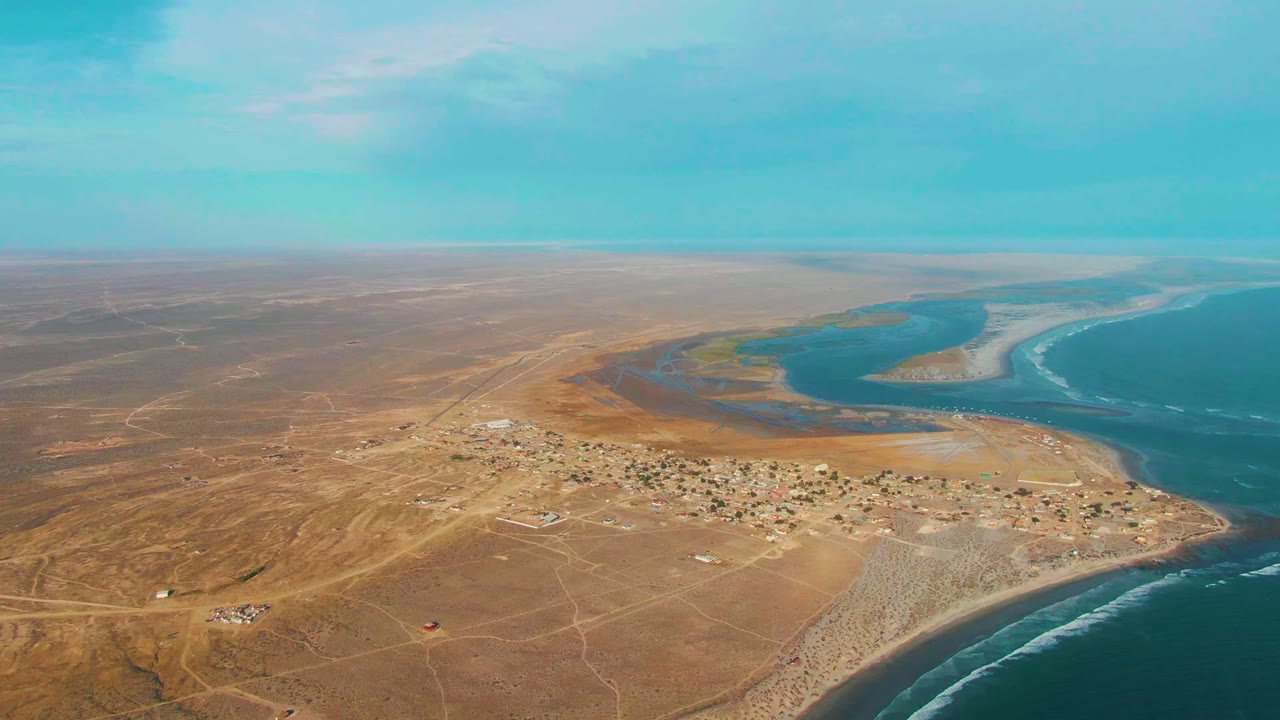 Wide aerial over a sandy coastal settlement with lagoon channels and open ocean.
