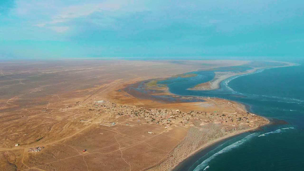 Aerial frame over La Bocana, Baja California Sur, showing a small desert village near a river mouth with blue ocean to the right and no visible people.