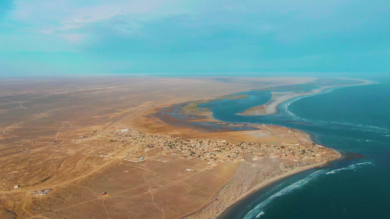 Wide aerial coast view with village, desert flats, river mouth, and ocean.