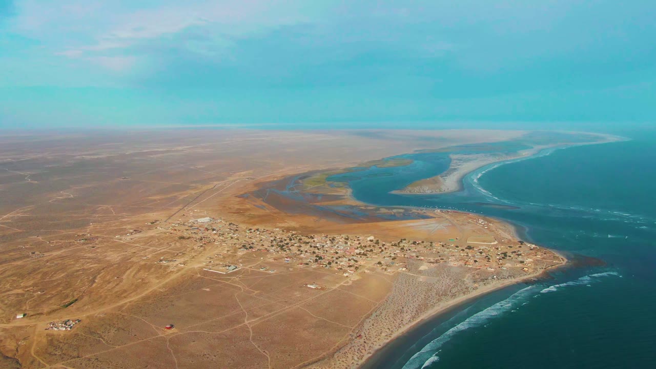 Aerial frame over the La Bocana coastal settlement, with tan desert foreground, a curving river mouth behind the village, blue ocean along the right side, and no visible people.