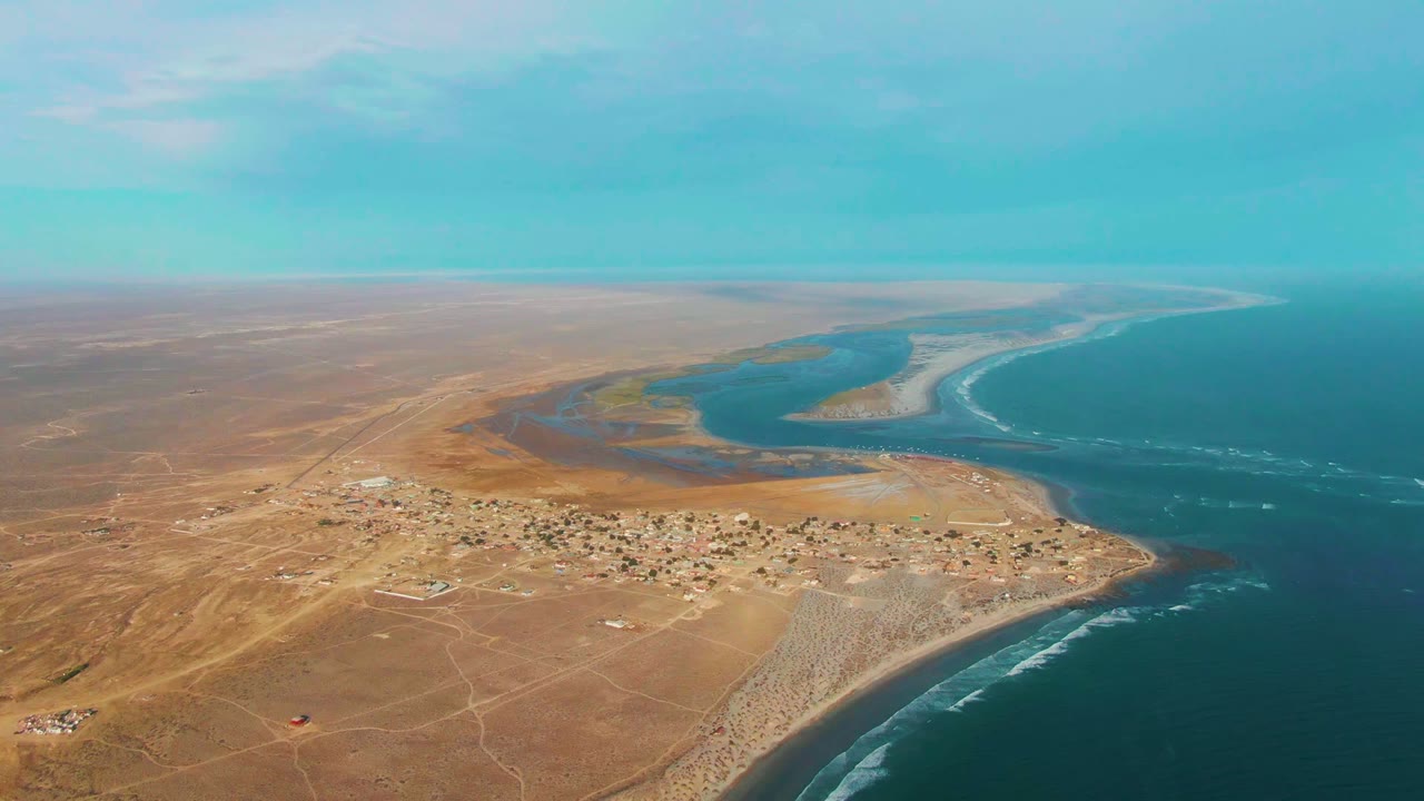 Aerial view over La Bocana, Baja California Sur showing tan desert terrain, a small coastal settlement, lagoon water, ocean surf, and no visible people at the start of the clip.