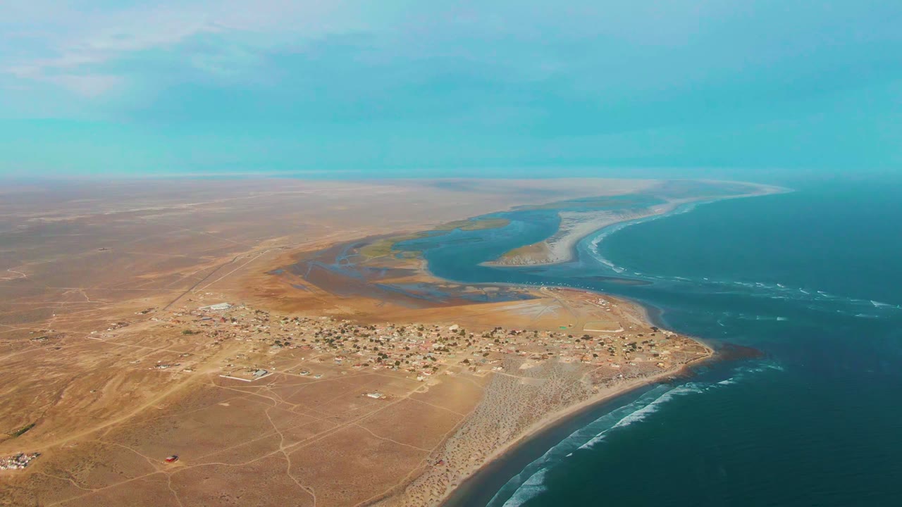 Aerial view of La Bocana showing arid foreground roads, a compact coastal settlement, lagoon water beyond, a long surf edge, and no visible people.