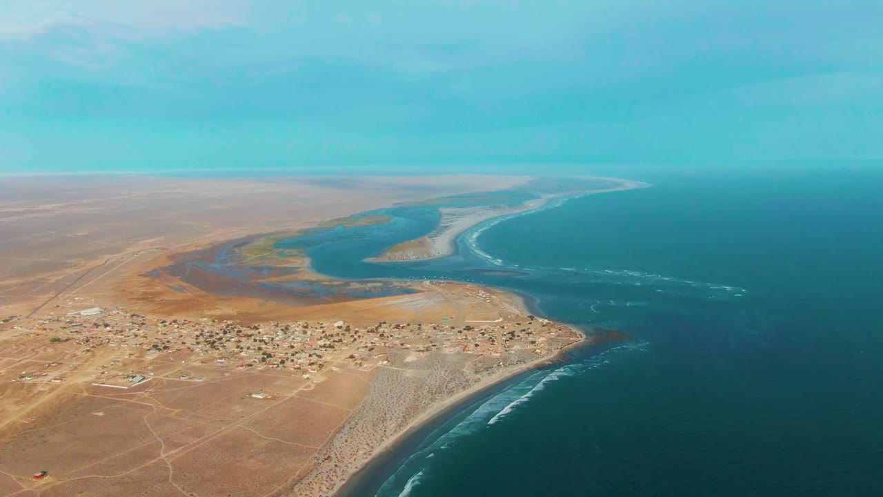 Opening aerial frame over La Bocana, Baja California Sur, showing tan desert land, a small coastal settlement, lagoon shallows, sandy beach, blue ocean, and no visible people.
