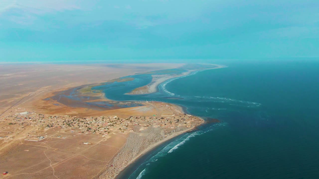 La Bocana aerial frame showing the lagoon curving through the coastal flats, a compact settlement near the beach, white surf lines on the ocean edge, and no visible people.