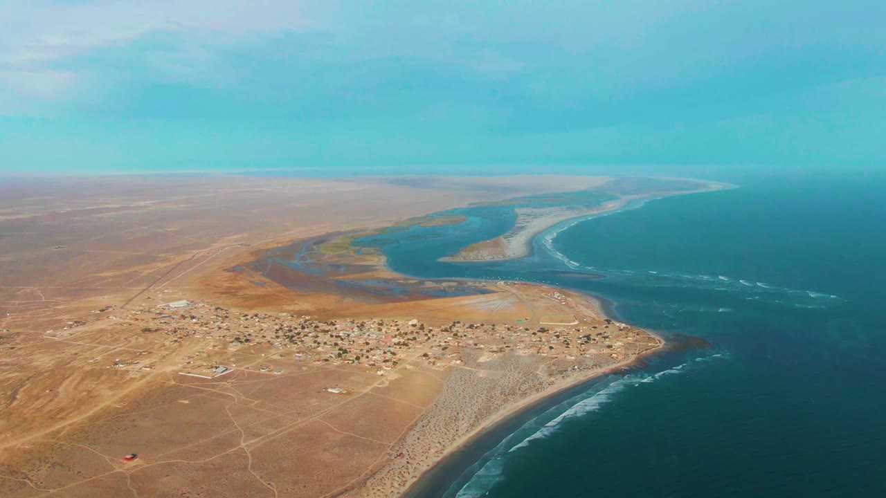 Opening aerial frame over La Bocana, Baja California Sur, showing a small desert settlement between dry land, a blue lagoon, and ocean surf, with no visible people.
