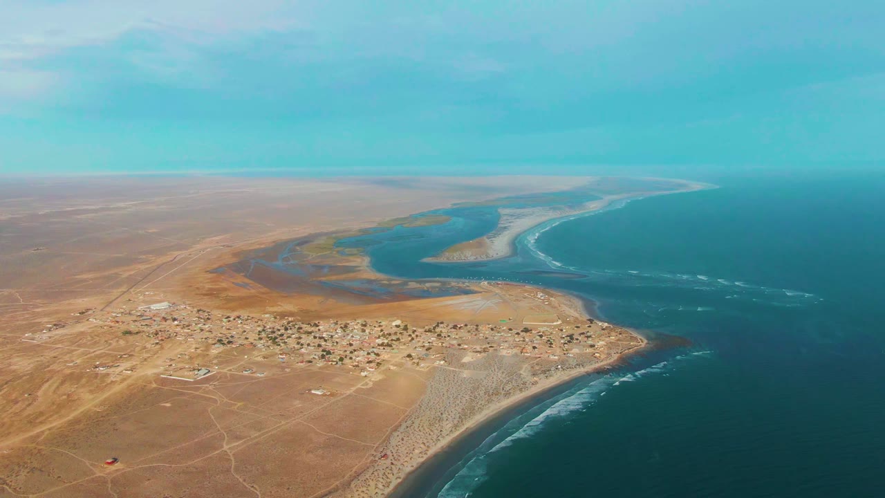 High drone view of La Bocana showing dry brown terrain, clustered buildings near the coast, lagoon channels behind the shore, and white surf marks on the ocean, with no visible people.