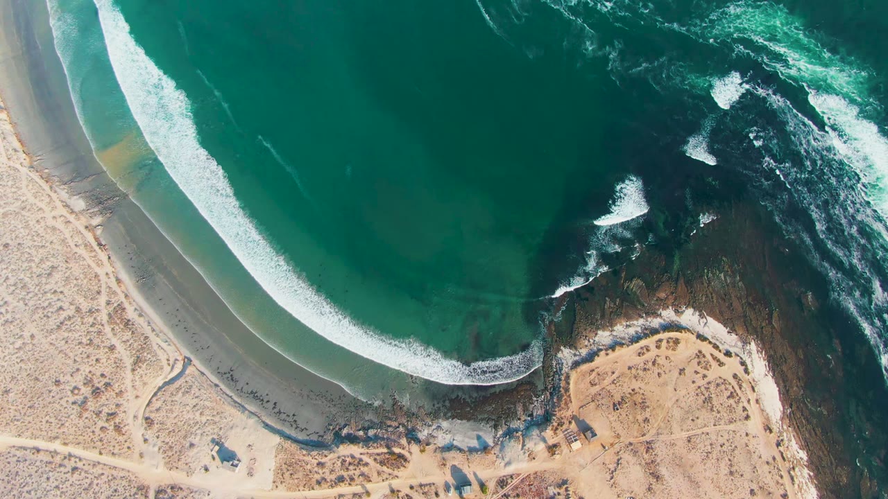 Aerial view over La Bocana, Baja California Sur, where a curved band of white surf meets tan sand beside dark coastal rock, with no visible people.