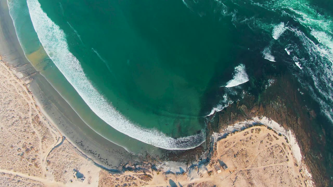 Top-down La Bocana shoreline view with turquoise water wrapping toward a sandy curve and breaking against dark rock on the right, with no visible people.