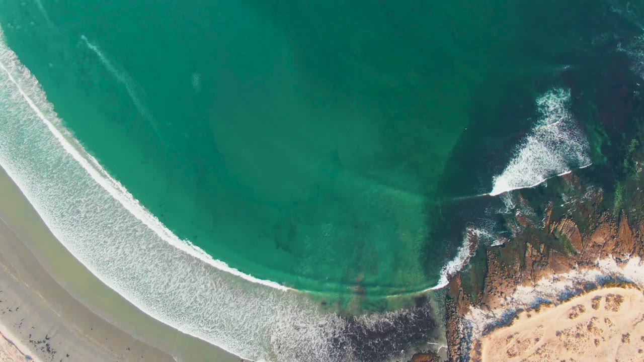 Aerial frame over La Bocana, Baja California Sur, with turquoise water, a curved white surf line, sandy beach, rust-colored rocks, and no visible people.