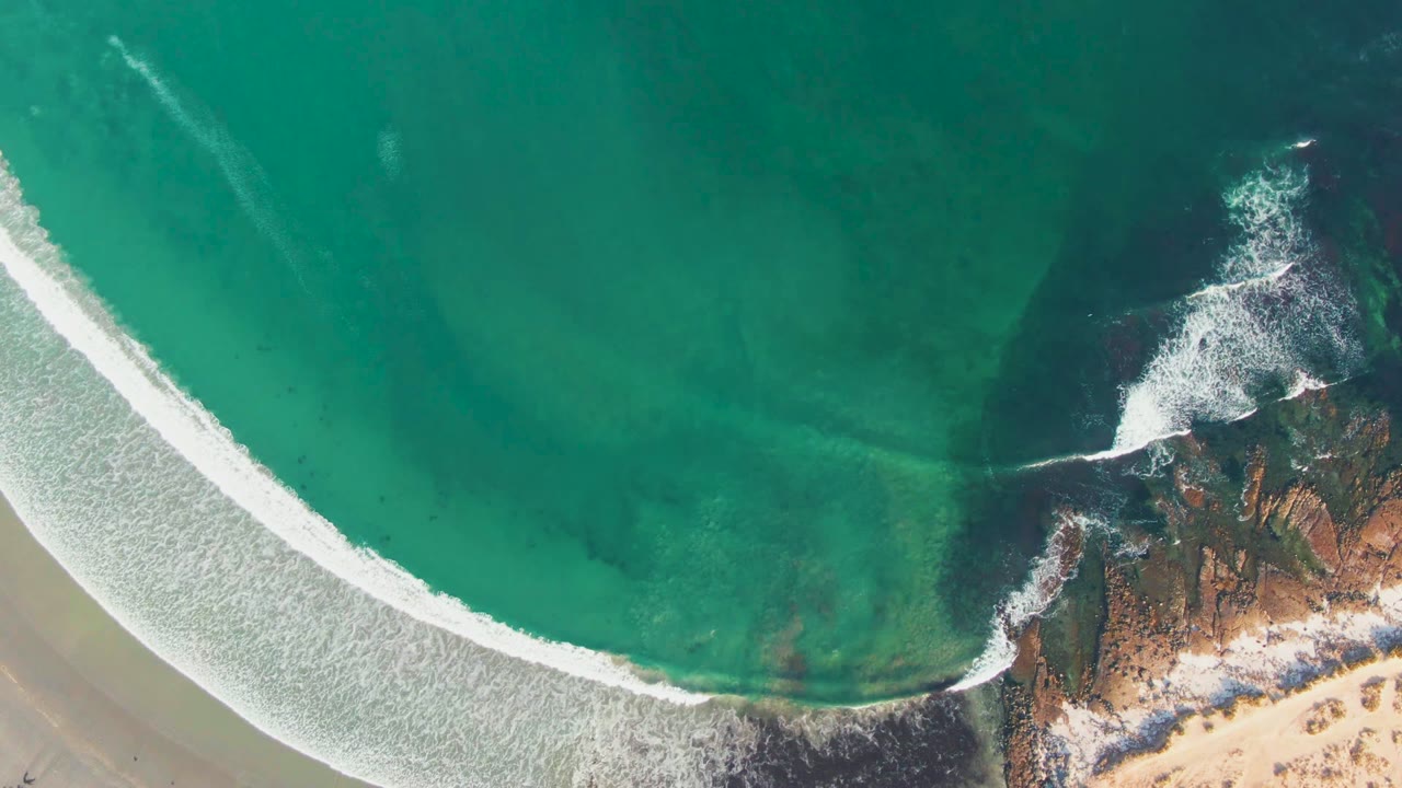 Aerial frame over La Bocana with surf foam spread wider across the shallows, rust-toned rocks at the lower right, pale sand below, and no visible people.
