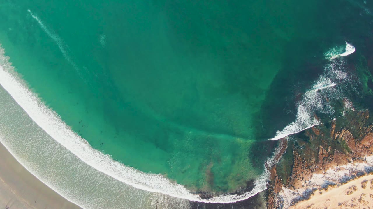 Overhead view in La Bocana, Baja California Sur, Mexico, with turquoise water, a white foam curve, sandy beach, rocky outcrops at right, and no visible people.